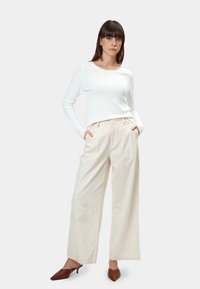 Weißer Langarmpullover kombiniert mit hellbeigen, weit geschnittenen Hosen mit Faltenwurf und strukturiertem Finish. Braune spitze Absatzschuhe.
