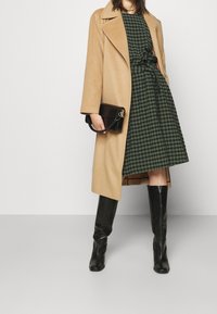 Abrigo de mezcla de lana de camello, vestido de cuadros verde y negro con cintura anudada, bolso de cuero negro y botas de cuero negro hasta la rodilla.