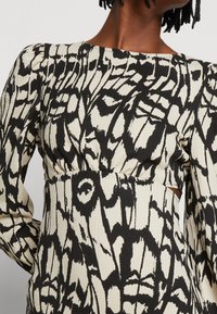 Robe à motifs noir et crème avec des manches longues, tissu texturé, taille cintrée et encolure ronde. Présente un design abstrait audacieux.