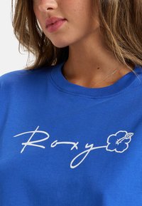 Camiseta de algodón azul con cuello redondo, que presenta el texto en cursiva blanca "Roxy" y un gráfico de flor de hibisco. Textura suave, diseño informal.