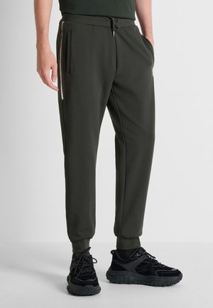 Uomo che indossa pantaloni da jogging verde scuro con dettaglio a striscia laterale e scarpe da ginnastica nere, in piedi con una mano in tasca.
