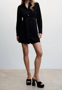 Robe noire à boutons avec manches longues, deux poches poitrine, taille cintrée et ceinture en tissu. Portée avec des mules noires à plateforme.