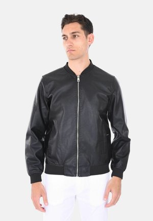 Uomo che indossa una giacca di pelle nera con zip e pantaloni bianchi, in piedi contro uno sfondo chiaro uniforme, che guarda di lato.