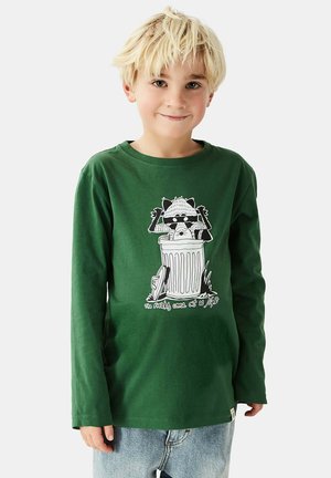 Blond child wearing a green long-sleeve shirt with a raccoon popping out of a trash can graphic and light blue jeans.