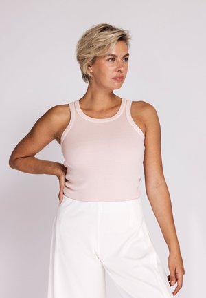Woman with short blonde hair wearing a light pink sleeveless ribbed top and white high-waisted wide-leg pants, posing with hand on hip.