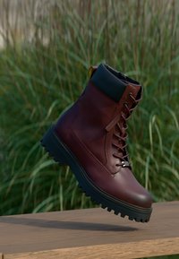 Καφέ δερμάτινο μποτάκι με κορδόνια και μαύρη επένδυση γύρω από τον αστράγαλο, παχύ μαύρο πέλμα με αυλακώσεις, αιωρούμενο πάνω από ξύλινη επιφάνεια με φόντο πράσινο γρασίδι.