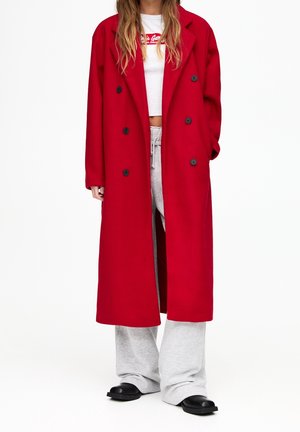 Persona con un abrigo largo rojo de doble botonadura, camiseta blanca corta, pantalones grises de chándal y zapatos negros sobre un fondo blanco.