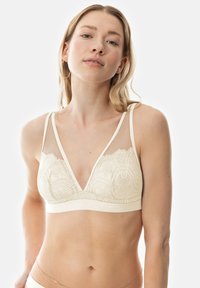Bralette beige clair avec panneaux en mesh transparent, motif en dentelle florale, décolleté plongeant et bretelles fines ajustables. Tissu doux et texturé.