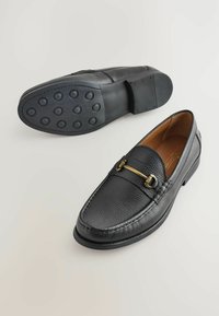 Mocasines de cuero negro con acabado texturizado, acentos de hardware dorado, suela de goma redonda con grips circulares y un diseño elegante.