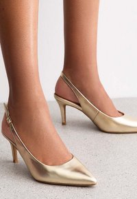 Des talons slingback dorés avec un bout pointu et un talon fin. Le matériau a une finition métallique lisse, comportant une sangle arrière pour un ajustement sécurisé.