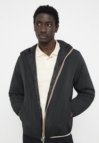 Chaqueta negra acolchada con capucha, que presenta un cierre de cremallera en contraste en naranja y plata, bolsillos laterales y una textura exterior suave.