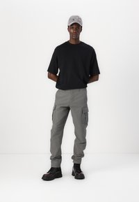 Tricou negru cu mânecă scurtă, pantaloni cargo gri cu buzunare laterale și cizme negre cu talpă groasă. O șapcă gri cu detalii brodate colorate.