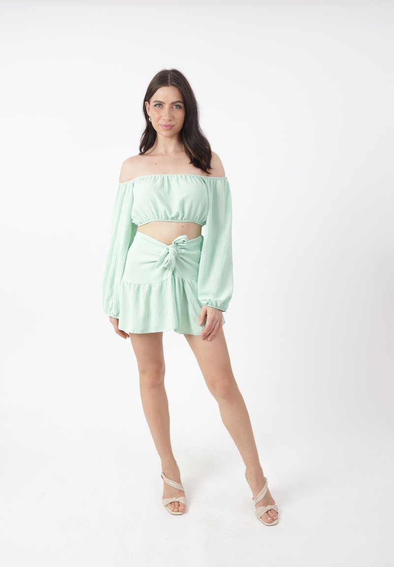 Mujer joven de pie con un top corto de tirantes caídos en verde menta y una falda atada a juego, con sandalias de tiras beige, sobre un fondo blanco.