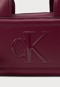 Burgundivörös bőr kézitáska, melyen dombornyomott "CK" logó található, sima felszínnel és kontrasztos varrással a széleken.