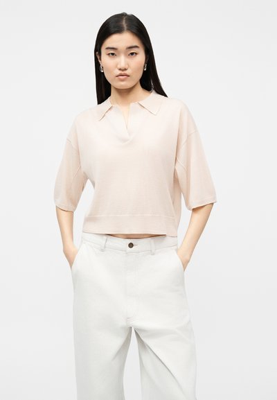 Νεαρή γυναίκα με ίσια μαύρα μαλλιά φοράει ανοιχτό μπεζ κοντομάνικο πουκάμισο με γιακά και λευκό ψηλόμεσο παντελόνι, με τα χέρια στις τσέπες.