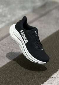 Baskets de course noires avec une tige texturée, une épaisse semelle blanche et un branding "HOKA" bien visible. Possède des lacets et des courbes douces dans son design.