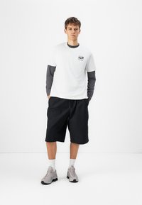 White t-shirt layered over a gray long-sleeve shirt, paired with black shorts and gray sneakers, featuring a logo on the chest.