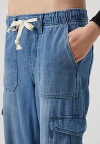 Ljusblå denim cargo-byxor med elastisk midja och en beige dragsko. Har sidofickor och stickade detaljer.