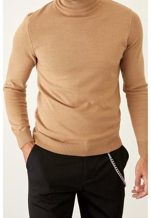 Homme portant un pull col roulé camel ajusté avec des poignets et un ourlet côtelés, et un pantalon noir avec un accessoire chaîne argentée.