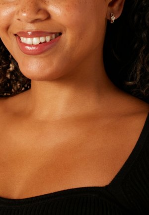 Gros plan d'une personne souriante portant de petites boucles d'oreilles argentées. Le teint est chaud, avec des taches de rousseur visibles, et le haut est noir avec une texture côtelée.