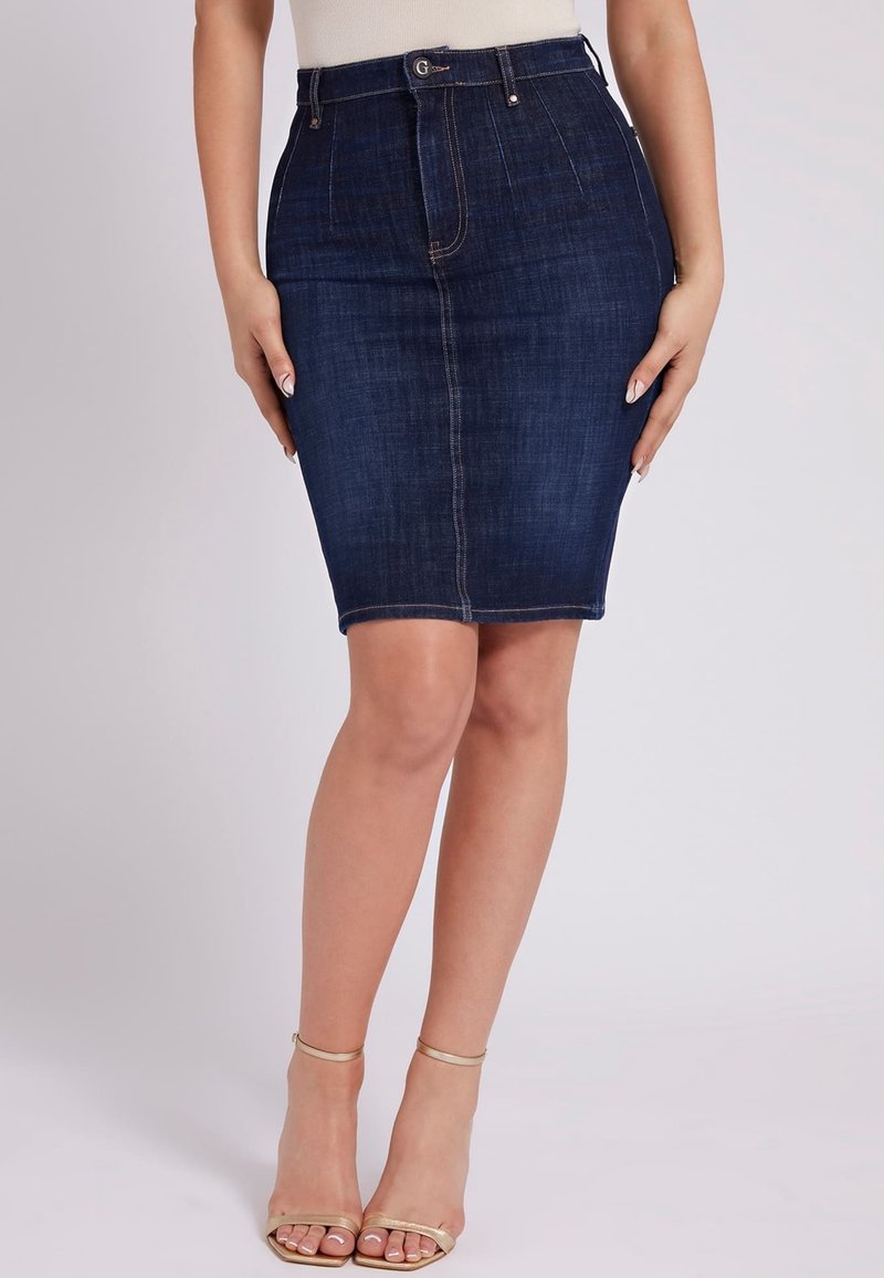 Jupe crayon en denim bleu foncé portée par une femme, associée à des talons à bride dorée à la cheville et un haut clair rentré.