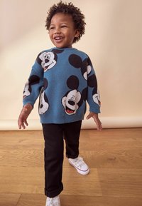 Maglione blu a maglia con grandi volti di Topolino, abbinato a pantaloni neri e sneaker bianche. Sfondo in legno.