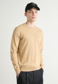 Beige kötött pulóver hosszú ujjakkal, kerek nyakkal és bordás mandzsettával. Fekete nadrággal viselve, amelyen egy kis kék logó található a mellkason.