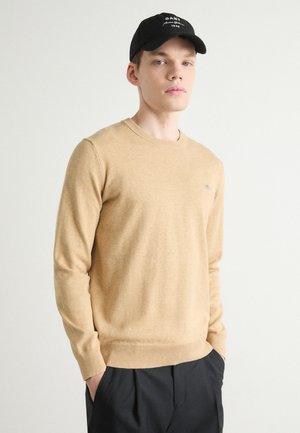 Beige Strickpullover mit langen Ärmeln, Rundhalsausschnitt und Rippbündchen. Getragen mit schwarzen Hosen, die ein kleines blaues Logo auf der Brust haben.