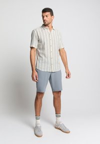 Kortarmig, lichtgekleurde shirt met verticale strepen, gecombineerd met grijze shorts. Model draagt grijze sneakers met witte en gestreepte sokken.