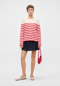 Sarkans un krēmkrāsas svītrots garām piedurknēm krekls, savienots ar tumši zilu džinsus svārku. Modelis tur mazu sarkanu somiņu, valkājot beigu krāsas ievelkamās kurpes.