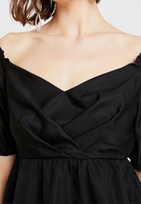 Černé šaty s odhalenými rameny a výstřihem do V, s nařasenými balónovými rukávy a splývavou sukní. Hladký materiál s jemným leskem.
