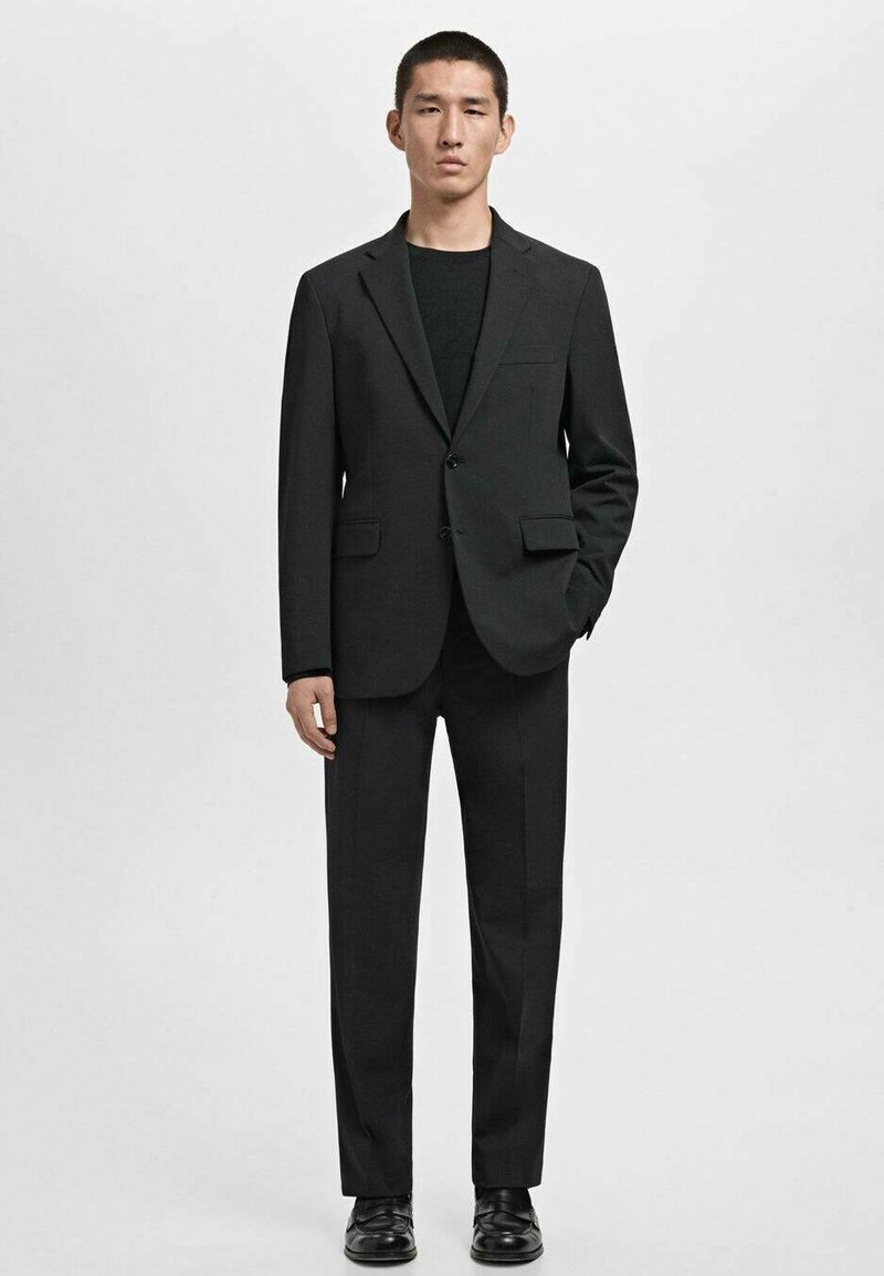 Traje negro con un blazer estructurado, cierre de un botón en el frente y dos bolsillos delanteros, combinado con pantalones a juego y zapatos negros.