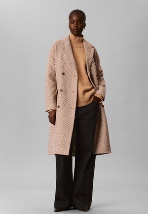 Femme portant un manteau beige texturé à double boutonnage sur un pull à col roulé beige avec un pantalon noir à jambes larges, sur un fond gris uni.