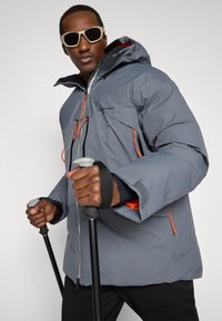 Šedá izolovaná bunda s oranžovými akcenty, předními kapsami a kapucí. Model drží trekingové hole a nosí sluneční brýle. Nepromokavý materiál.
