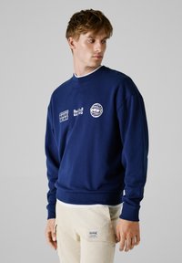 Sweatshirt bleu marine à col ras du cou, avec des logos imprimés blancs sur la poitrine et la manche. Fabriqué en coton mélangé doux et texturé.