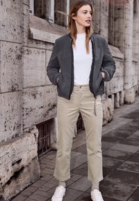 Jeune femme portant une veste grise, une chemise blanche, un pantalon beige et des baskets blanches, debout sur un trottoir en ville, les mains dans les poches, regardant vers la droite.