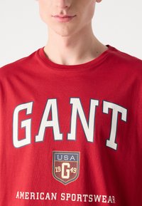 Tricou roșu din bumbac cu guler rotund, având text alb îndrăzneț "GANT" și "AMERICAN SPORTSWEAR," împreună cu un logo în formă de scut dedesubt.