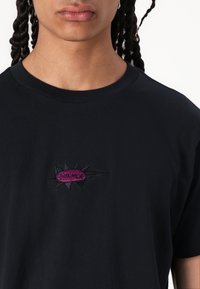 Svart bomullströja med en cirkulär broderad logotyp där ordet "FORMER" är i rosa, omgiven av svarta taggar. Standard design med rund hals.