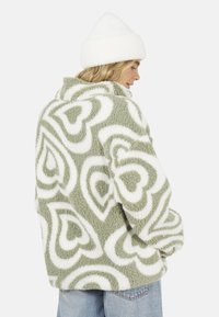 Fluffy groene jas met witte hartpatronen, met een hoge kraag en een ontspannen pasvorm. Gebreide witte beanie op het hoofd van het model.