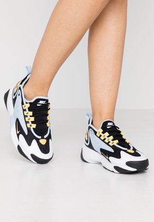 Nohy v černobílých a světle modrých teniskách Nike se zlatými akcenty a nápisem "ZOOM 2000" na tkaničkách stojí na světlé podlaze.