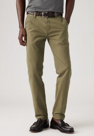 Uomo che indossa pantaloni chinos verde oliva, una camicia grigia infilata dentro con una cintura intrecciata in pelle marrone scuro e mocassini in pelle neri.