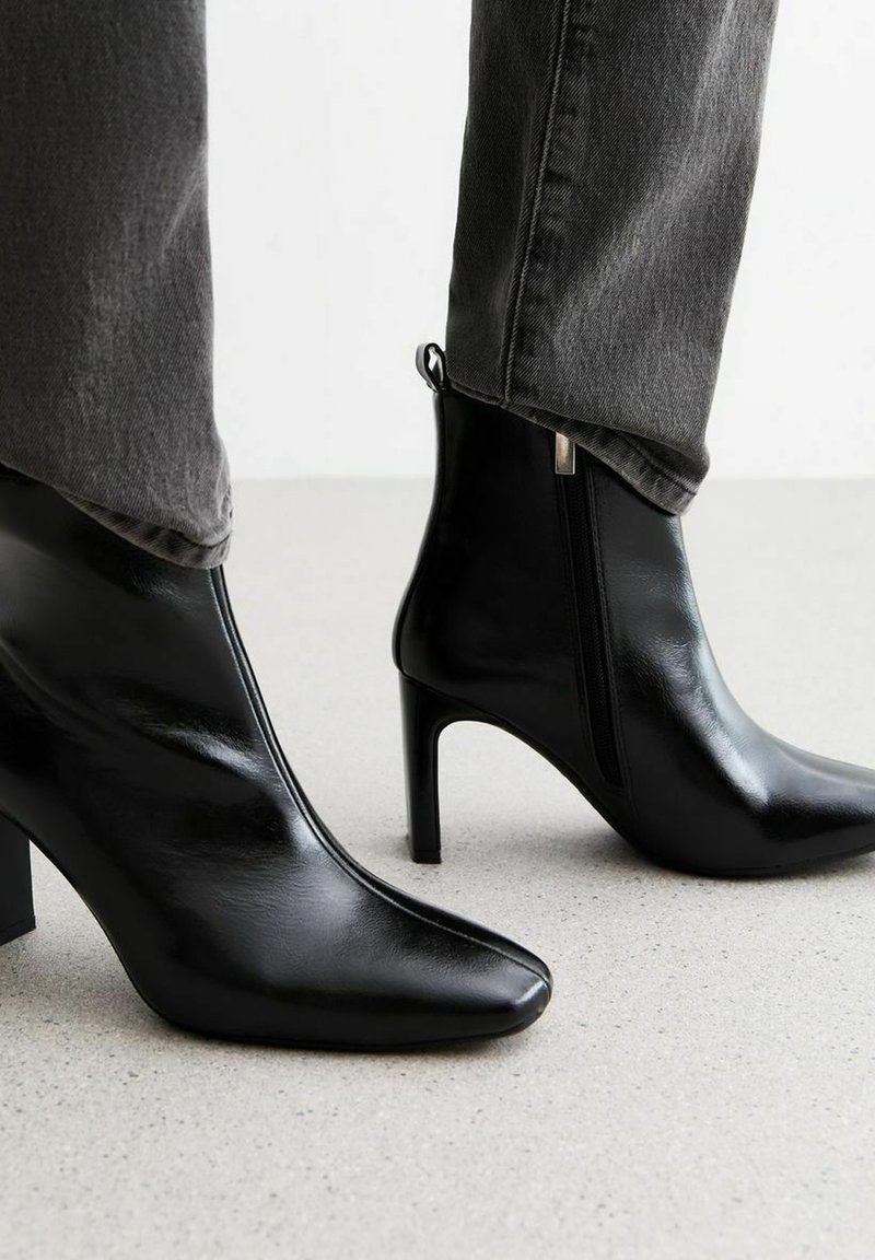Zwarte high-heeled enkellaarsjes met een spitse neus en glanzende afwerking. Voorzien van een rits aan de zijkant en een lus voor eenvoudig aan- en uittrekken. Geverfde grijze jeans zichtbaar.