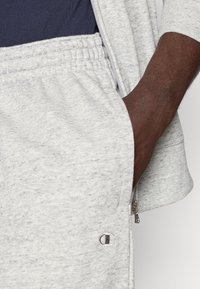 Ύφασμα φούτερ σε γκρι απόχρωση με υφή, με ελαστική μέση και πλευρική τσέπη με φερμουάρ. Διακριτική λεπτομέρεια λογότυπου στο πόδι.