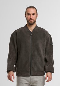 Giacca in pile grigia con colletto alto, chiusura frontale a zip e vestibilità comoda. Presenta una piccola patch con logo sulla manica sinistra e una texture morbida.