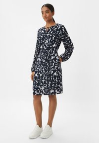 Robe bleu marine avec un motif abstrait blanc, manches longues, un nœud au niveau du col, et une taille cintrée. Associée à des baskets blanches.