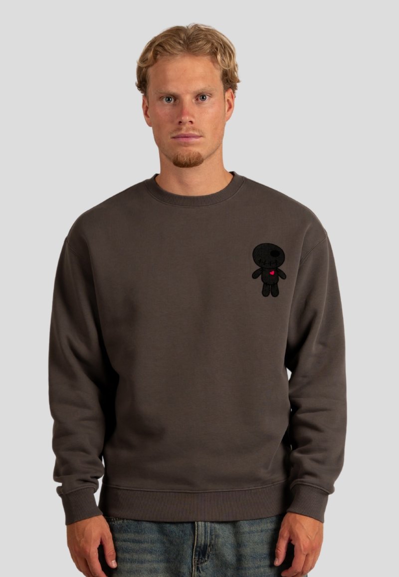 Felpa grigia con polsini e orlo a coste. Presenta un personaggio ricamato in nero sul lato sinistro del petto, con un dettaglio a cuore rosso.