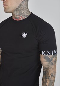 Melnā pieguļošā T-krekls ar balto izšūto logo uz krūtīm un ar joslām pie piedurknēm, kurā melnā fonā ir teksts "SikSilk" baltā krāsā.