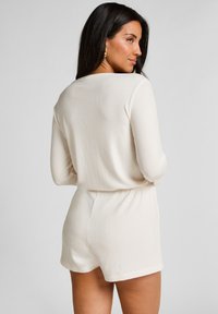 Jumpsuit de crema con textura de canalé, mangas largas, cinturilla elástica y pantalones cortos. Cuenta con un clásico escote redondo y una tela suave y texturizada.