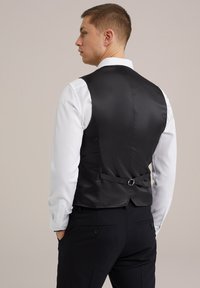 Vesta din satin negru cu o textură netedă, având o curea ajustabilă la spate. Purtați peste o cămașă albă.