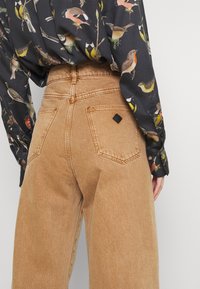 Bruna kordbyxor med hög midja och vida ben, med två bakfickor och en svart tygetikett nära den högra fickan.